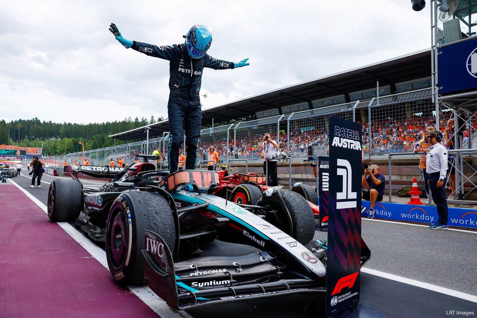 George Russell standing on top of his car celebrating his win at the 2024 Austrian Grand Prix in Parc Ferme, with pit lane and grandstands in the background.