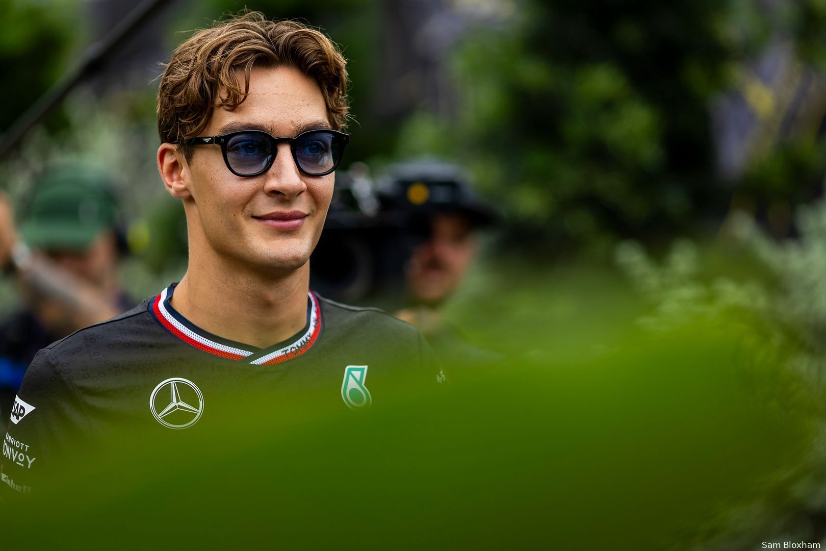 George Russell in Mercedes team clothes at the Singapore Grand Prix paddock, wearing glasses, with a camera crew in the background, shot from behind a green plant.