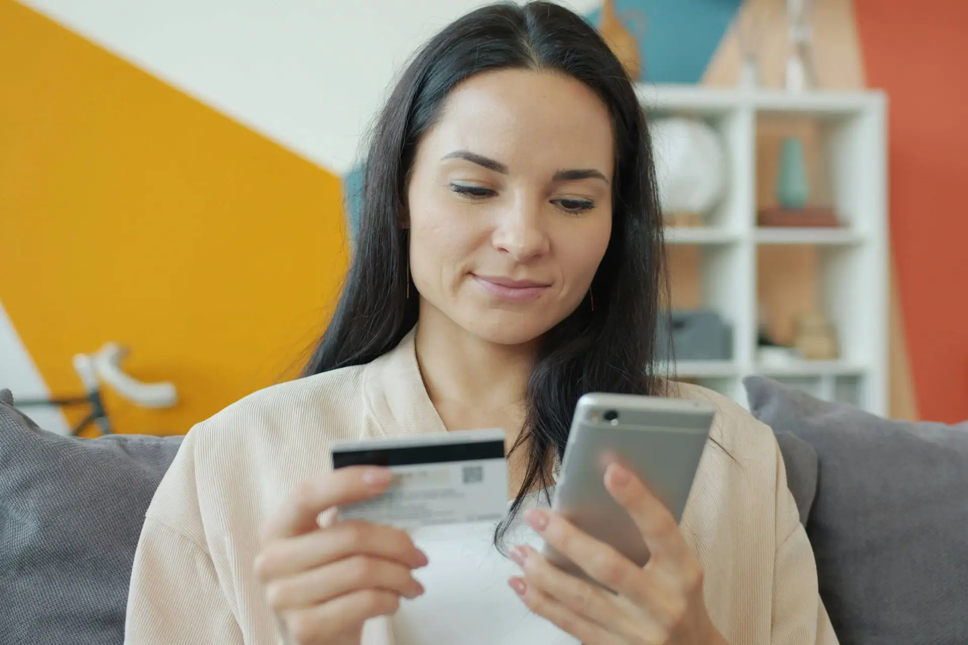 A woman sitting  on a gray sofa holding a credit card and looking at a smartphone.