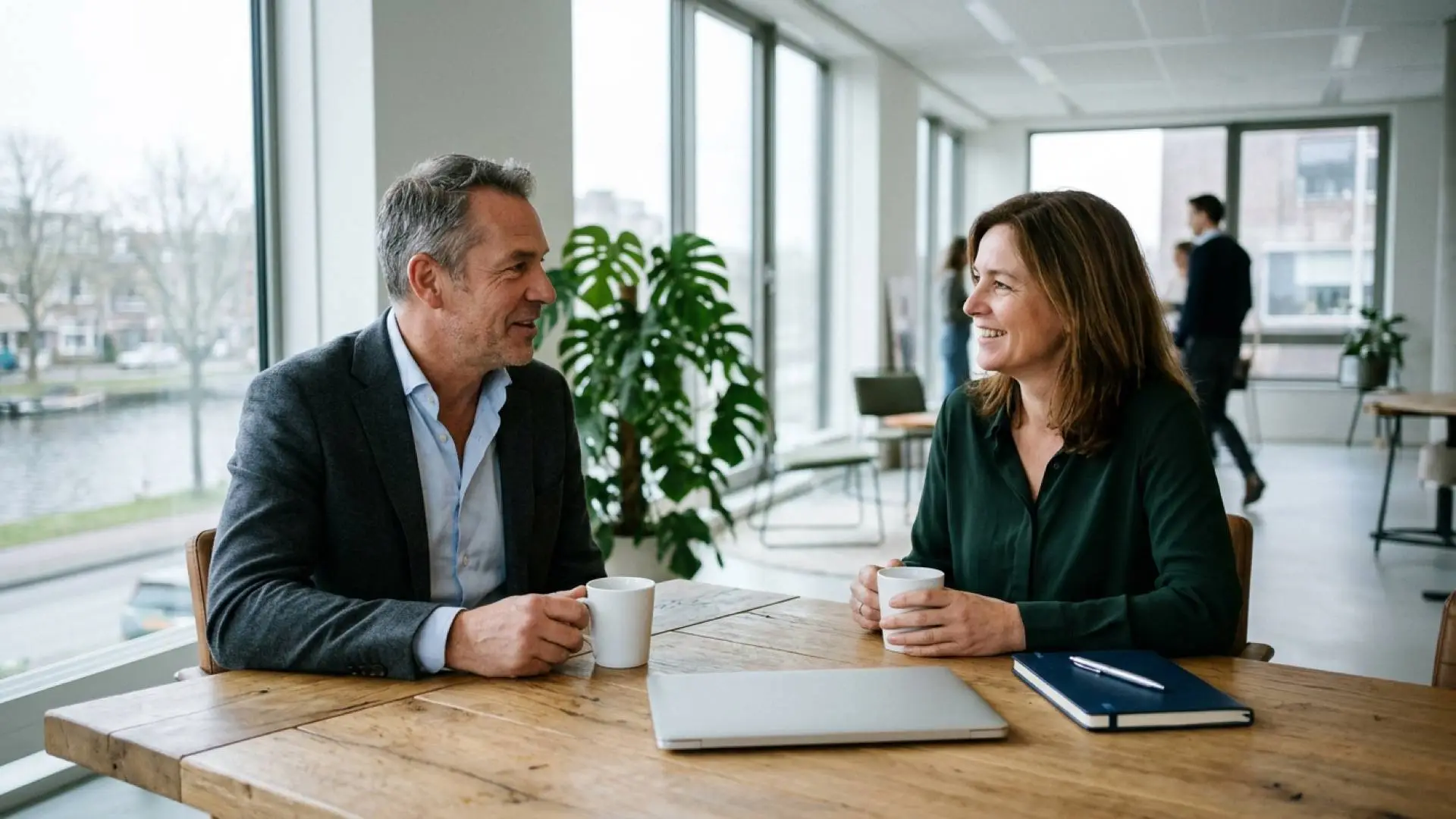 Nederlandse man en vrouw zitten in een lichte kantoorruimte aan een houten tafel met twee koffiemokken en een laptop