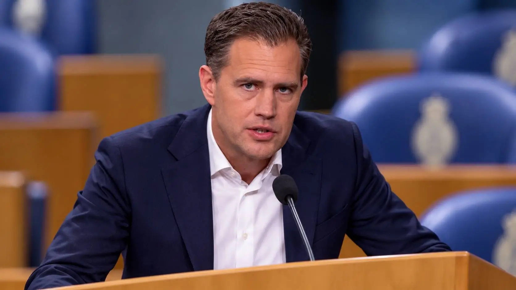 AI-illustratie van Eelco Heinen achter het spreekgestoelte in de Tweede Kamer, met blauwe Kamerbankjes op de achtergrond.