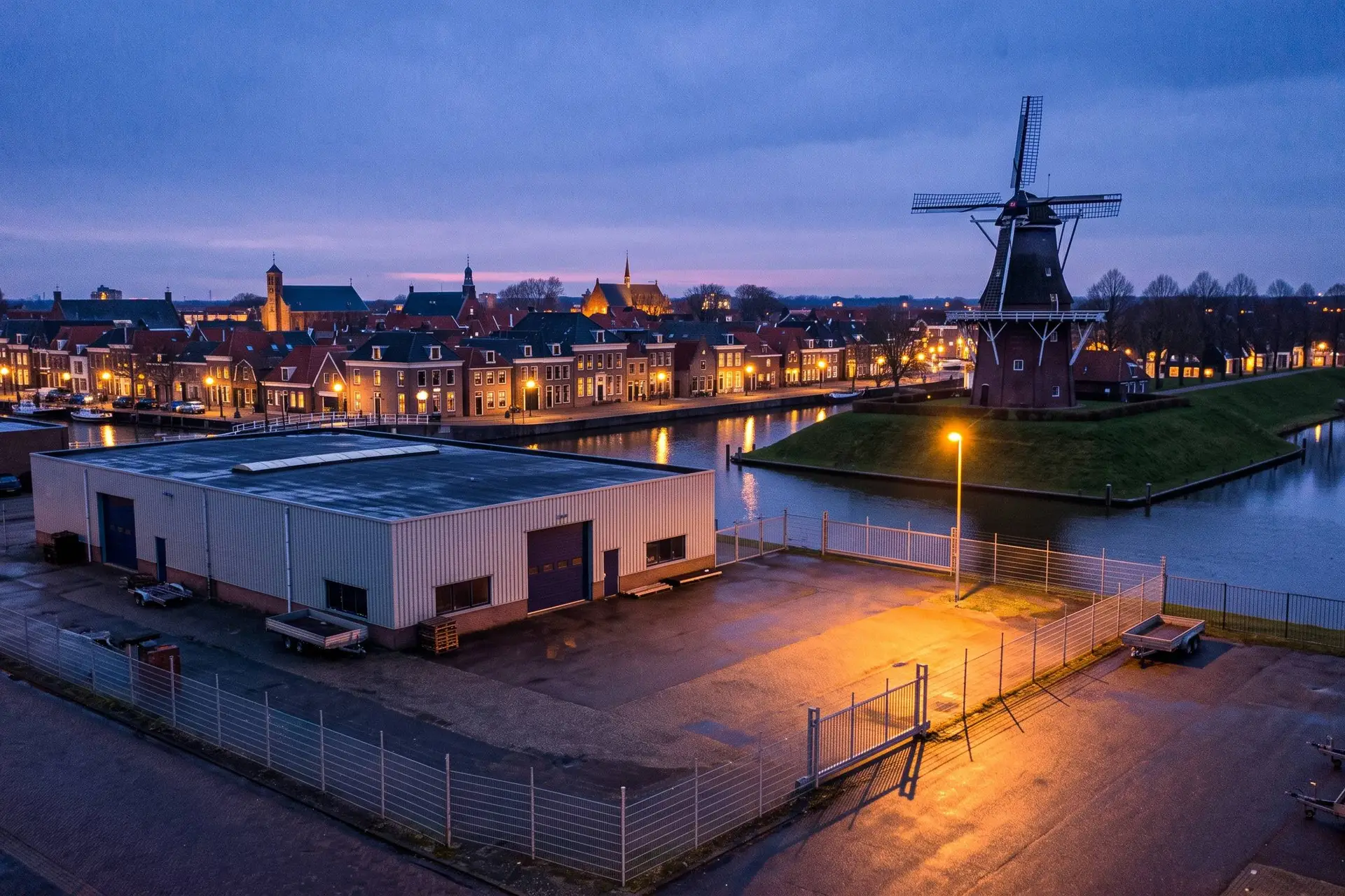 Leeg bedrijfsterrein met een laag gebouw aan de rand van een Friese stad met gracht en molen bij schemering