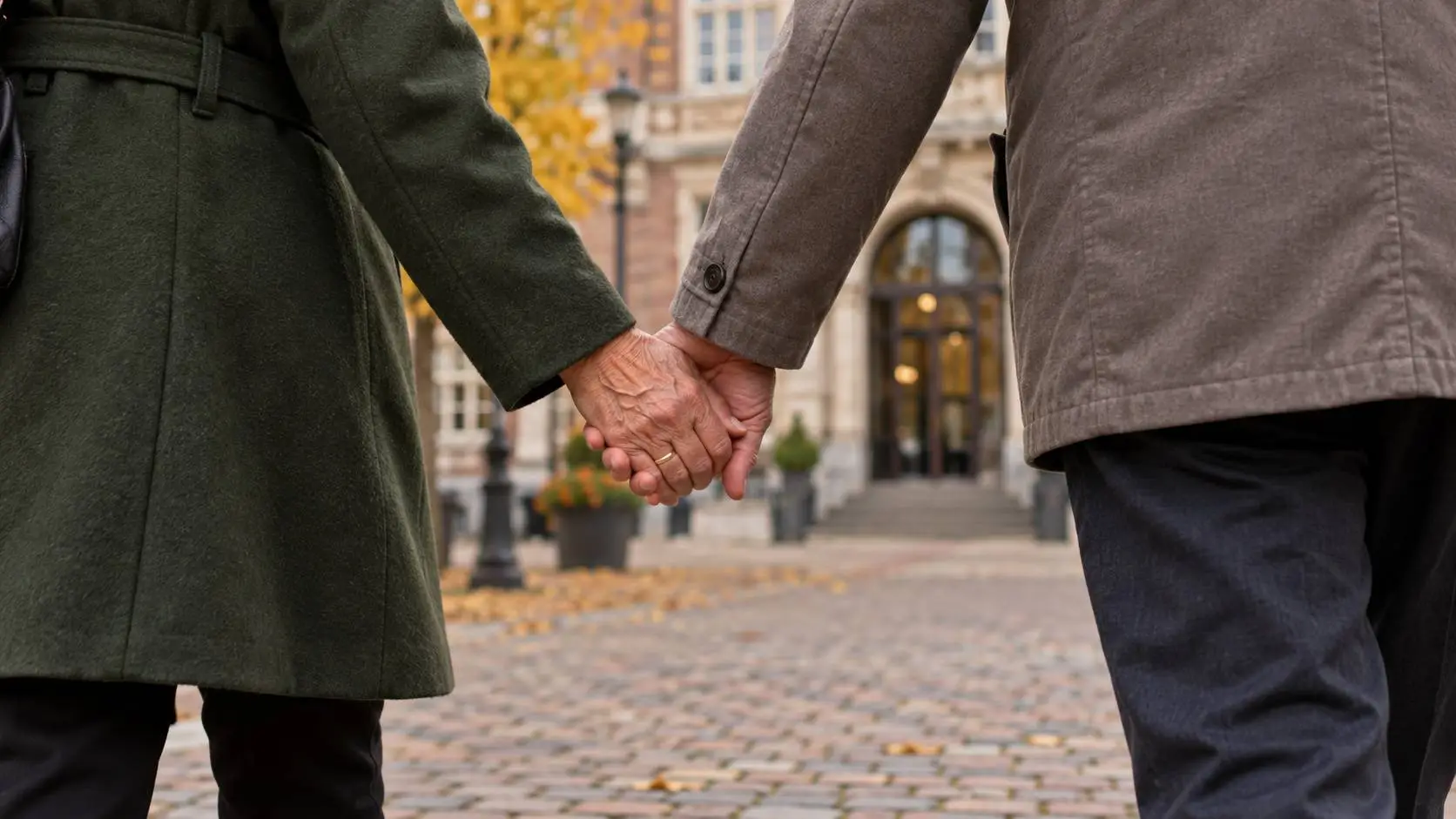 Twee oudere mensen die hand in hand over een plein bij een gemeentehuis lopen.