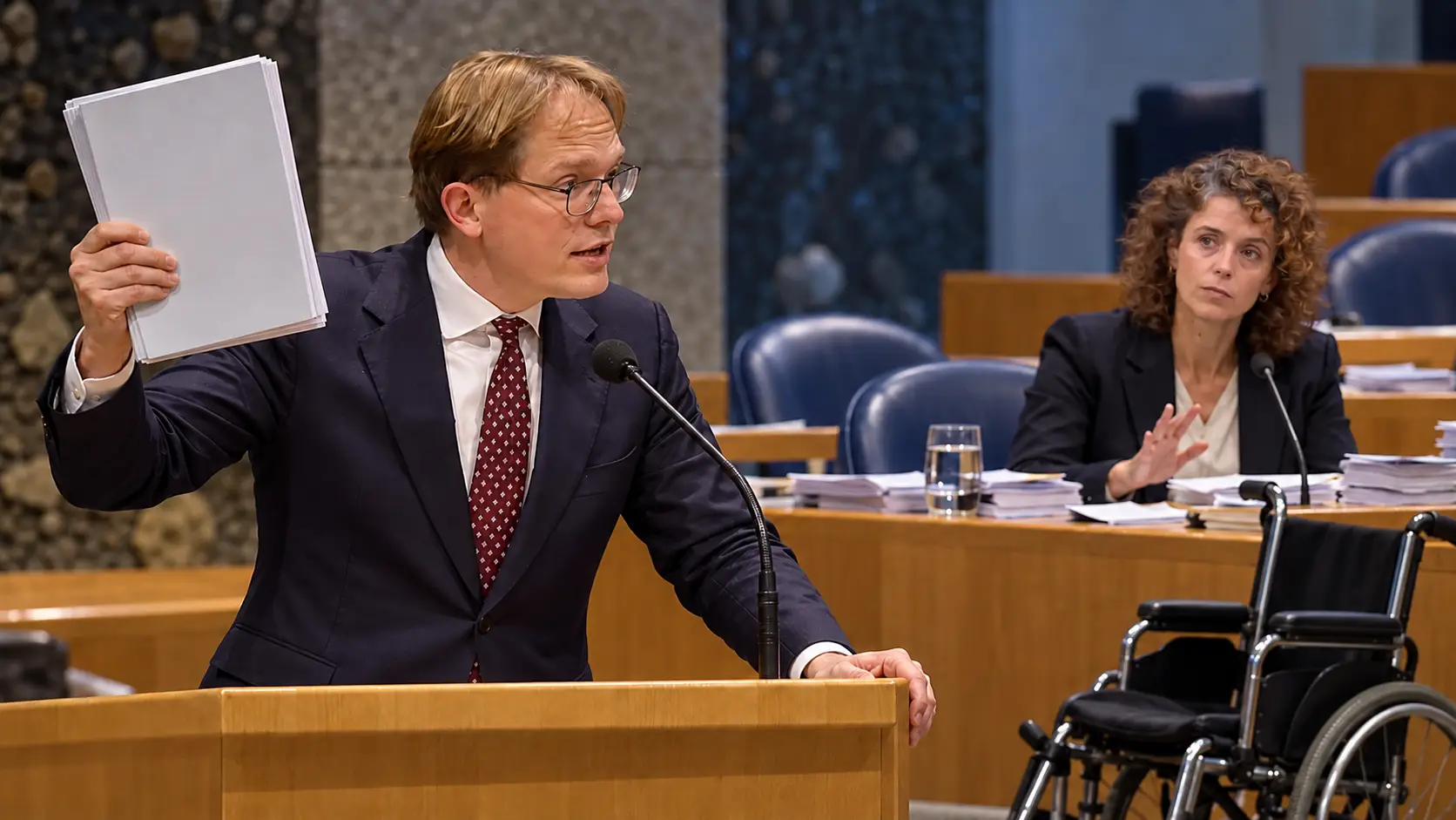 Illustratie van Pepijn van Houwelingen met een begrotingsmap, Sophie Hermans achter de regeringstafel en een lege rolstoel op de voorgrond.