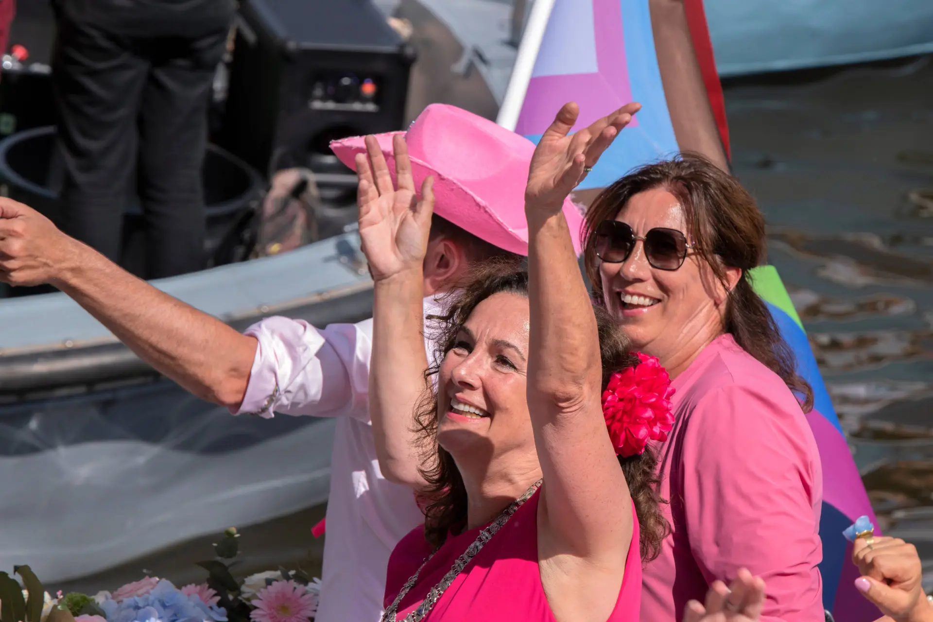 Femke Halsema zwaait lachend vanaf een boot met een regenboogvlag, in een roze shirt met een bloem in het haar
