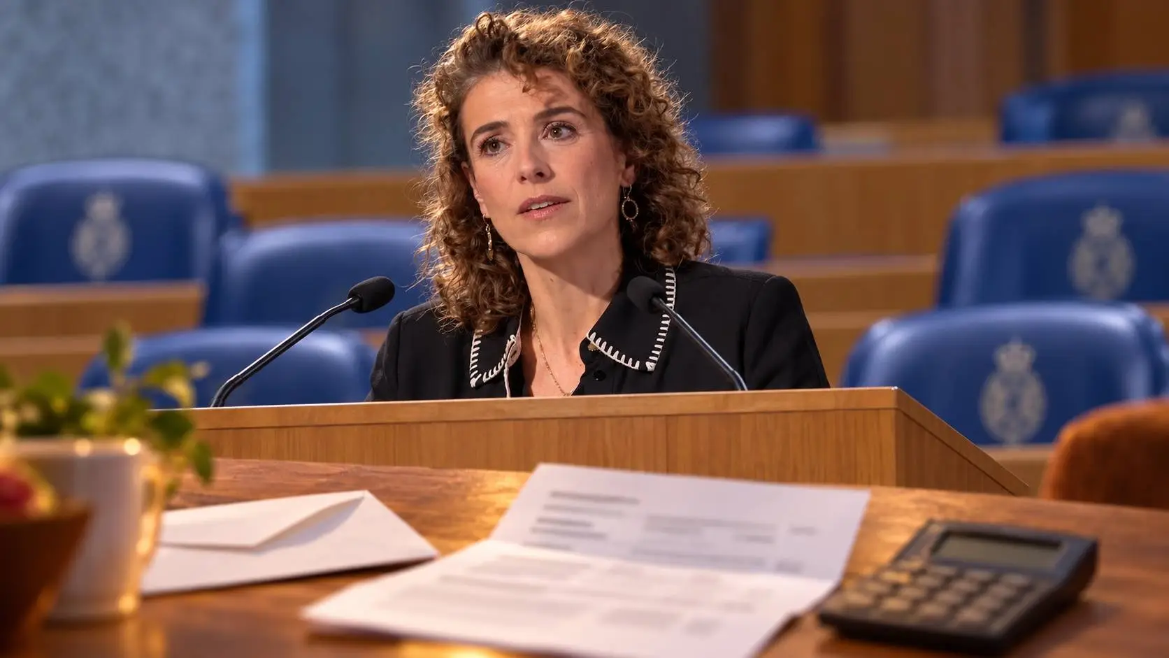 Illustratie van Sophie Hermans achter het spreekgestoelte in de Tweede Kamer, met op de voorgrond een zorgverzekeringsbrief en een rekenmachine op een keukentafel.