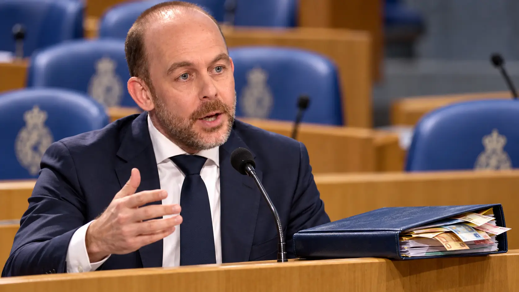 Illustratie van Bart van den Brink bij een spreekgestoelte in de Tweede Kamer, naast een begrotingsmap met bankbiljetten.