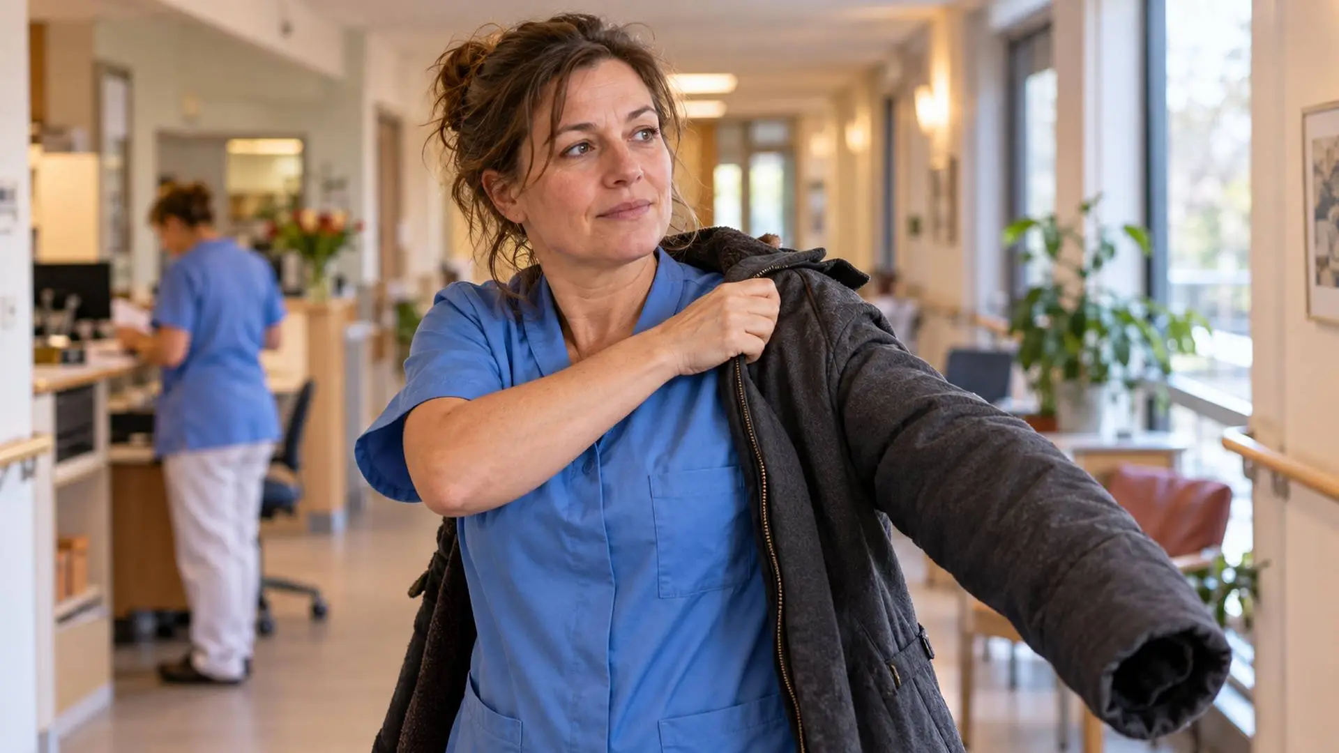 Zorgmedewerker trekt na haar dienst haar buitenjas aan in een lichte gang van een verpleeghuis.