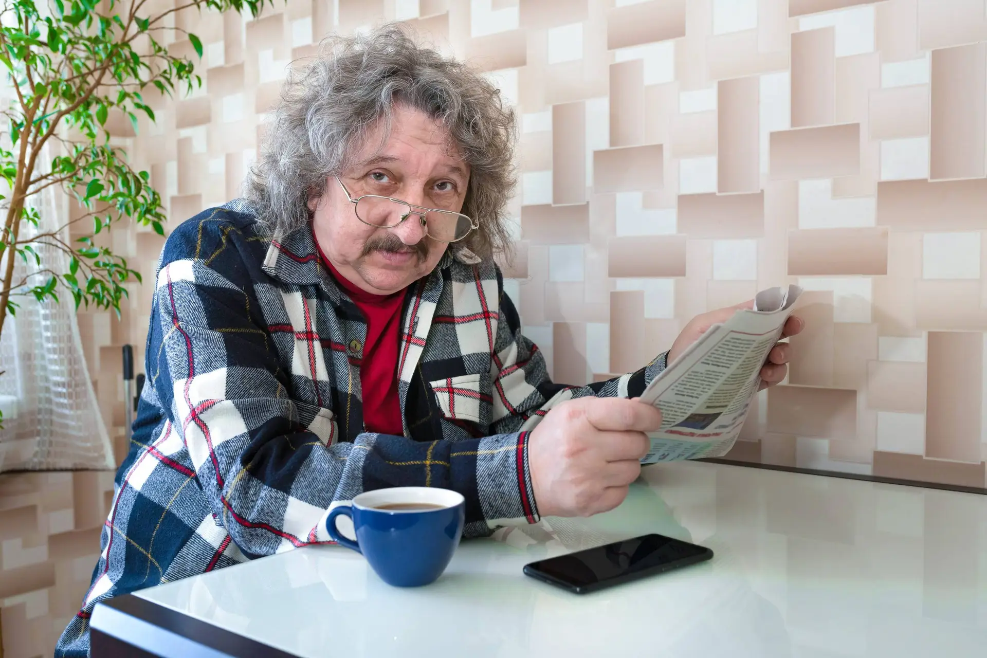 Een oudere 60-jarige man zit aan tafel met een kop koffie en een krant in zijn handen.