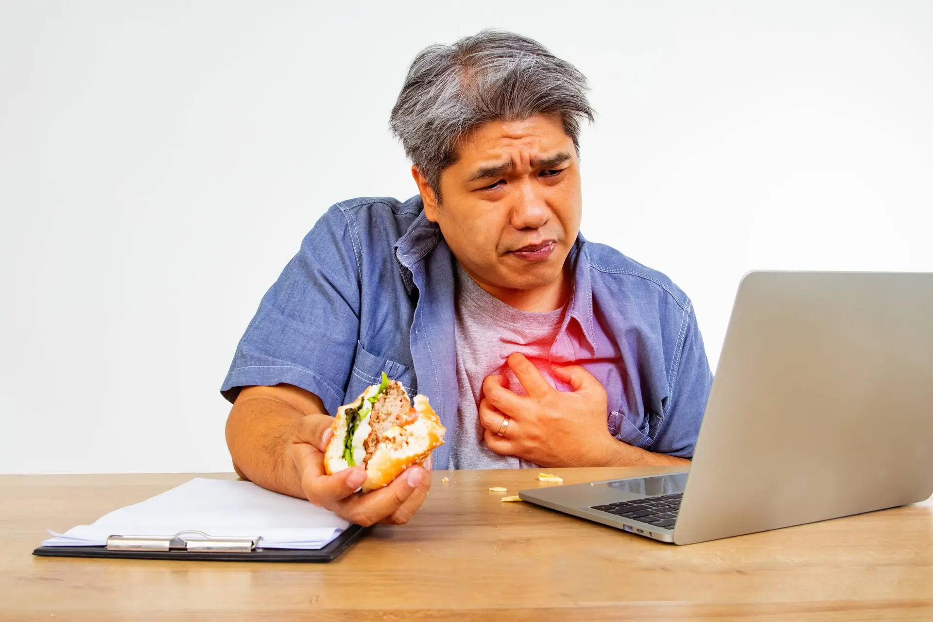 Deze man van middelbare leeftijd eet een hamburger tijdens het zitten op het werk, laptop, computer, heeft pijn op de borst, ongemakkelijke ademhaling, hartziekte, raakt zijn pijnlijke hart met zijn handen.