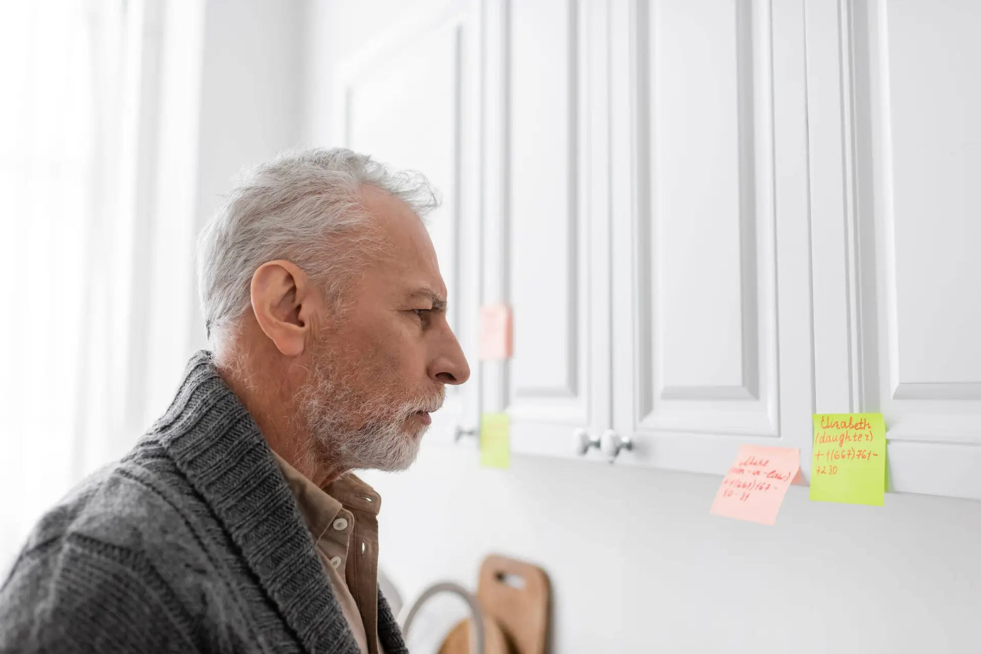 zijaanzicht van grijsharige man met alzheimer syndroom kijkend naar plakkerige notities met namen en telefoonnummers in de keuken