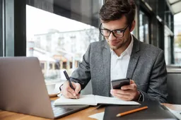 smiling-businessman-eyeglasses-sitting-by-table-cafe-with-laptop-computer-while-using-smartphone-writing-something