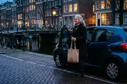 Vrouw van rond de zestig met boodschappentas stapt in de schemering uit haar auto langs een Amsterdamse gracht