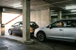Twee grijze auto's rijden vlak achter elkaar onder de opengaande slagboom van een parkeergarage door richting de uitrit.