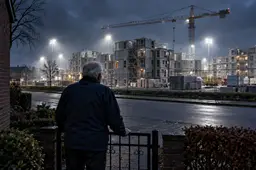 Man bij tuinhek kijkt in de schemering naar een fel verlichte bouwplaats met woonblokken, symbool voor het azc in Uden