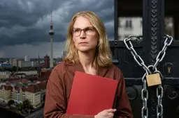 D66-minister Elanor Boekholt-O'Sullivan met een rode map bij een hek met ketting en hangslot, met Berlijn op de achtergrond