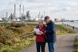 Ouder echtpaar leest een brief op een dijk met een raffinaderij op de achtergrond