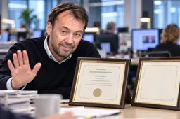 Satirische AI-illustratie van Pieter Klok met een afwerend handgebaar aan een redactietafel, naast een ingelijst diploma en een lege lijst.