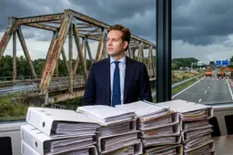 VVD-minister Vincent Karremans achter stapels aanbestedingsdossiers, met een roestige stalen brug en wegwerkzaamheden