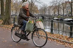 Oudere vrouw met een sjaal fietst met twee volle boodschappentassen langs een Nederlandse gracht in het najaar.