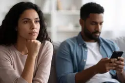 Een vrouw kijkt peinzend voor zich uit op de bank terwijl haar partner naast haar op zijn telefoon kijkt.