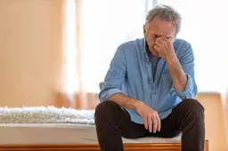 Een man van middelbare leeftijd zit ’s ochtends vermoeid op de rand van zijn bed.
