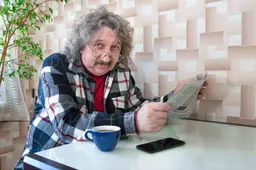 Een oudere 60-jarige man zit aan tafel met een kop koffie en een krant in zijn handen.