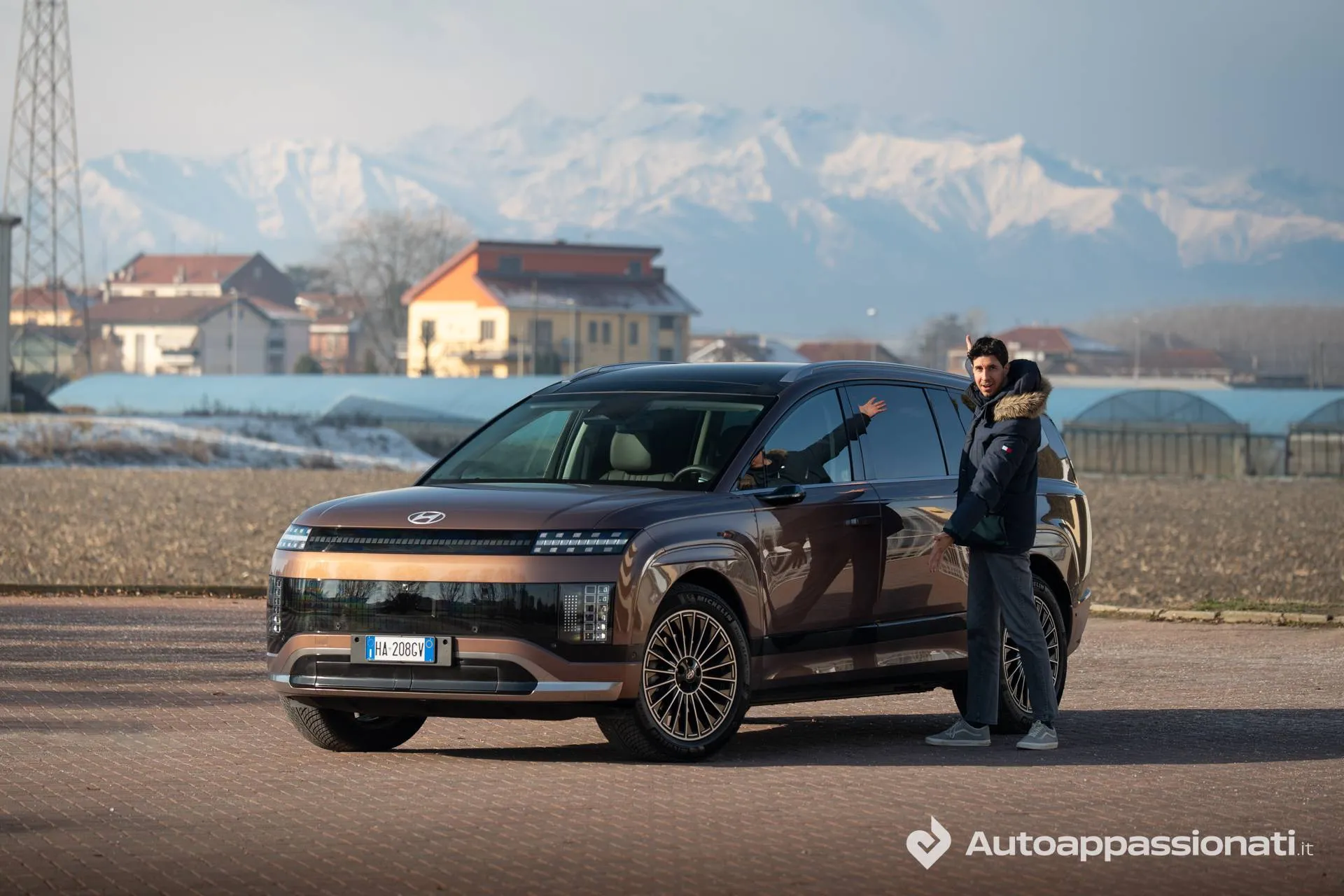 Esterni della nuova Hyundai IONIQ 9: il SUV elettrico color bronzo è ritratto di tre quarti anteriore con le montagne innevate sullo sfondo, mentre il tester ne indica le dimensioni imponenti.