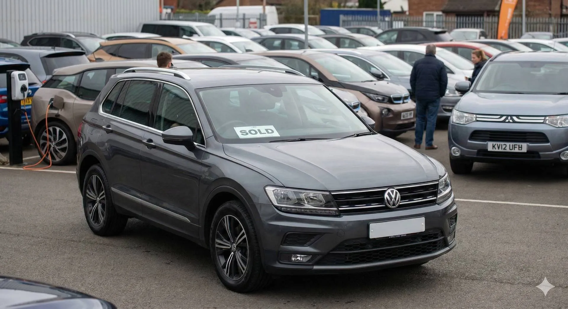 Una concessionaria di auto usate all'aperto con un SUV Volkswagen Tiguan grigio in primo piano che espone un cartello "VENDUTO" sul cruscotto. Sullo sfondo si notano diverse altre vetture di seconda mano.