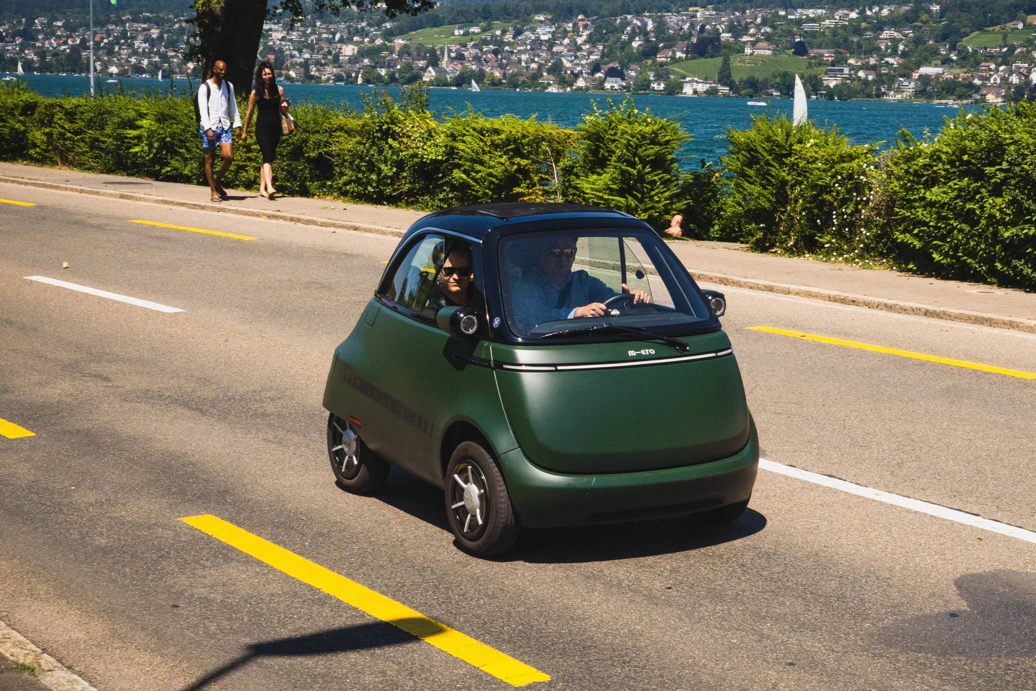 micro microlino di colore verde che percorre una strada con a bordo due persone. sullo sfondo due persone che passeggiano