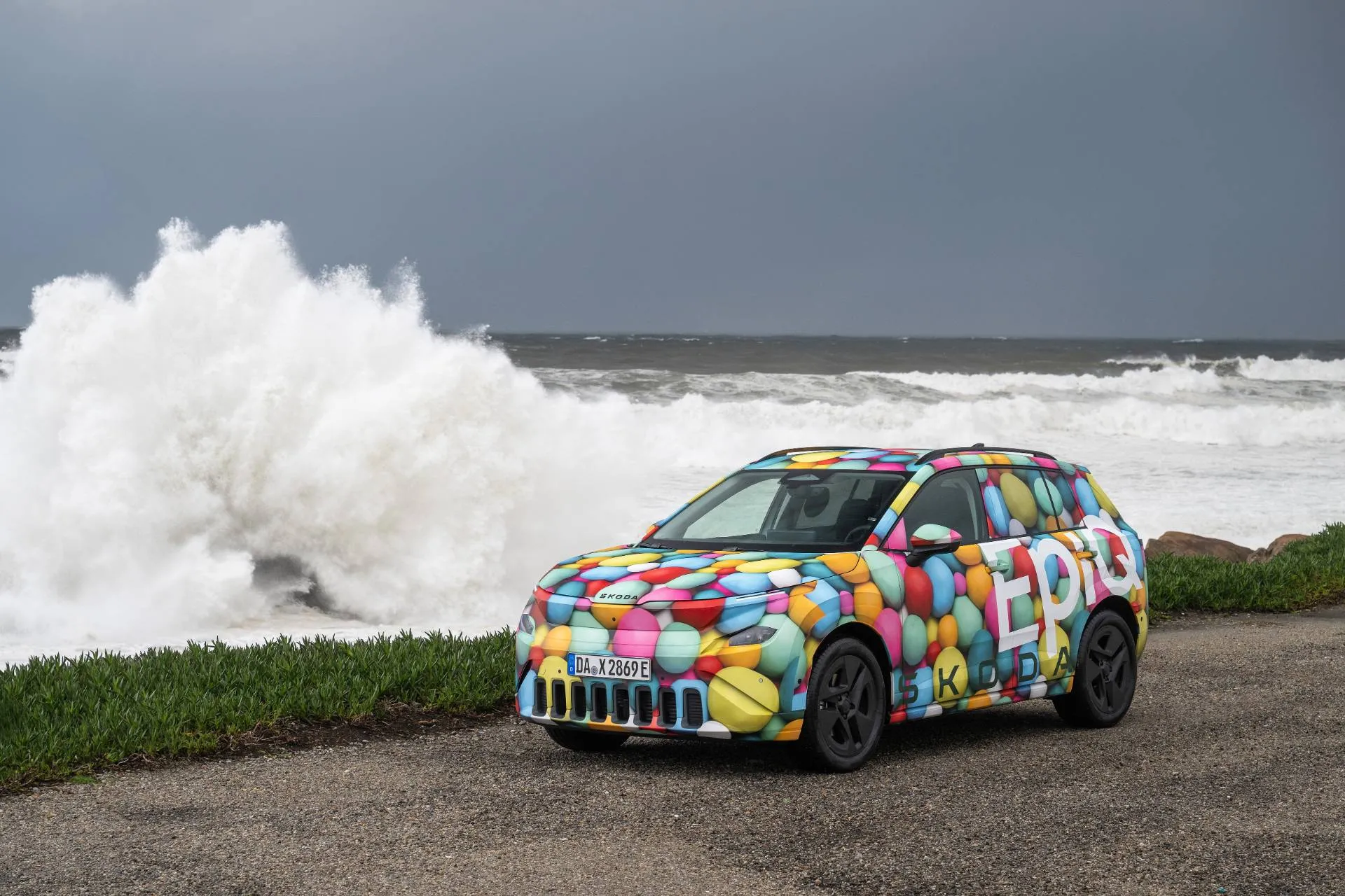 auto multicolore che mostra la nuova Skoda Epiq, crossover piccolo elettrico del marchio Skoda. Sullo sfondo il mare mosso con un'onda