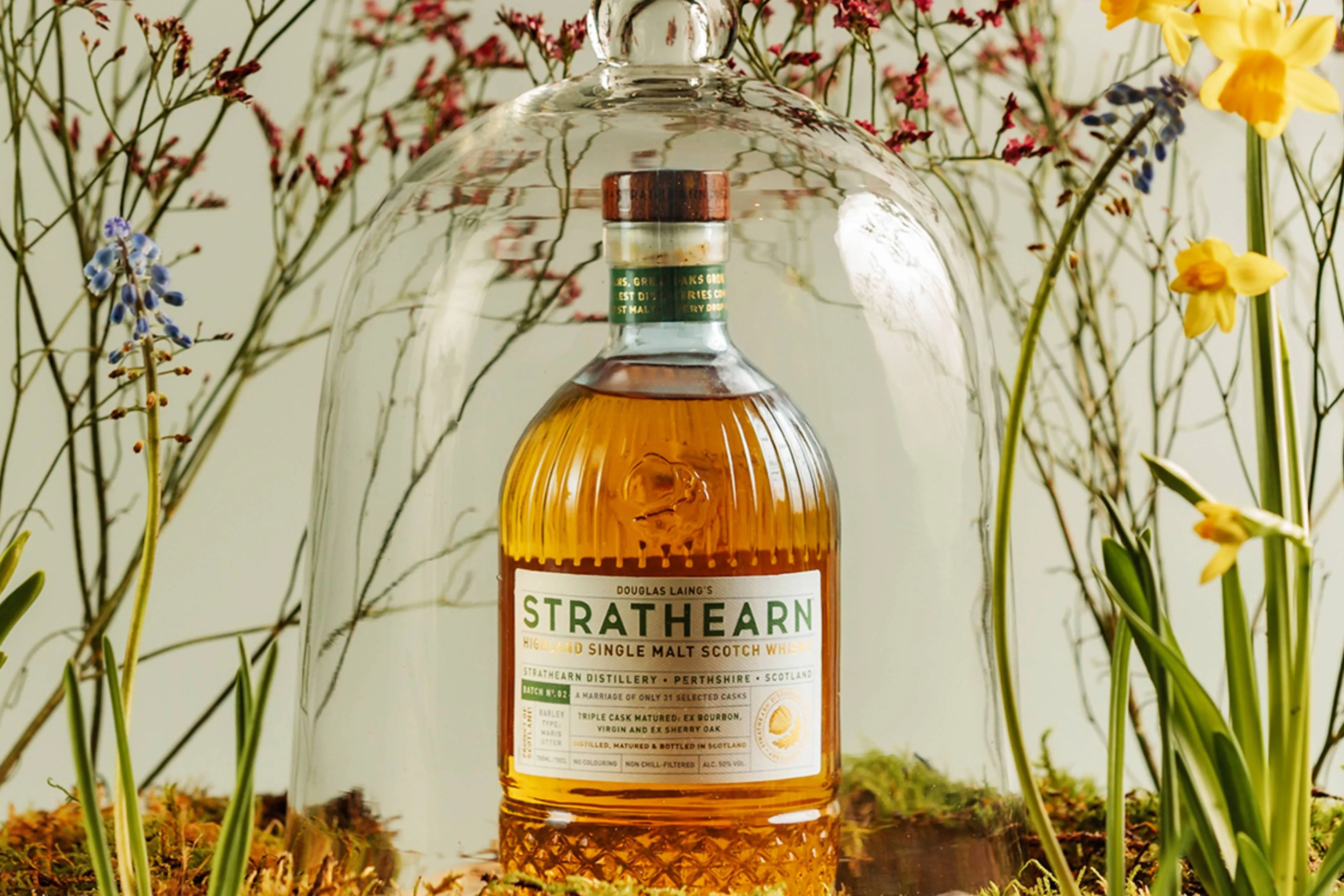 Een fles Strathearn Batch 2 whisky onder een stolp bij lentebloemen