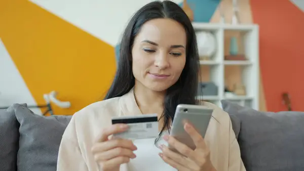 A woman sitting  on a gray sofa holding a credit card and looking at a smartphone.