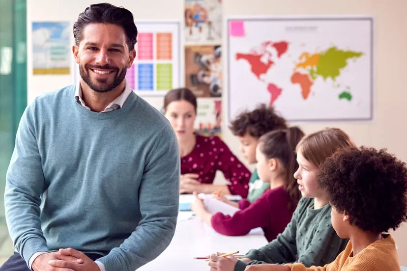 portrait smiling male elementary school teacher working desk classroom with students