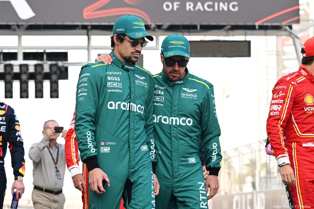 Fernando Alonso walks alongside Lance Stroll in team racing suits, with Alonso's hand resting on Stroll's shoulder, sharing a moment of camaraderie.