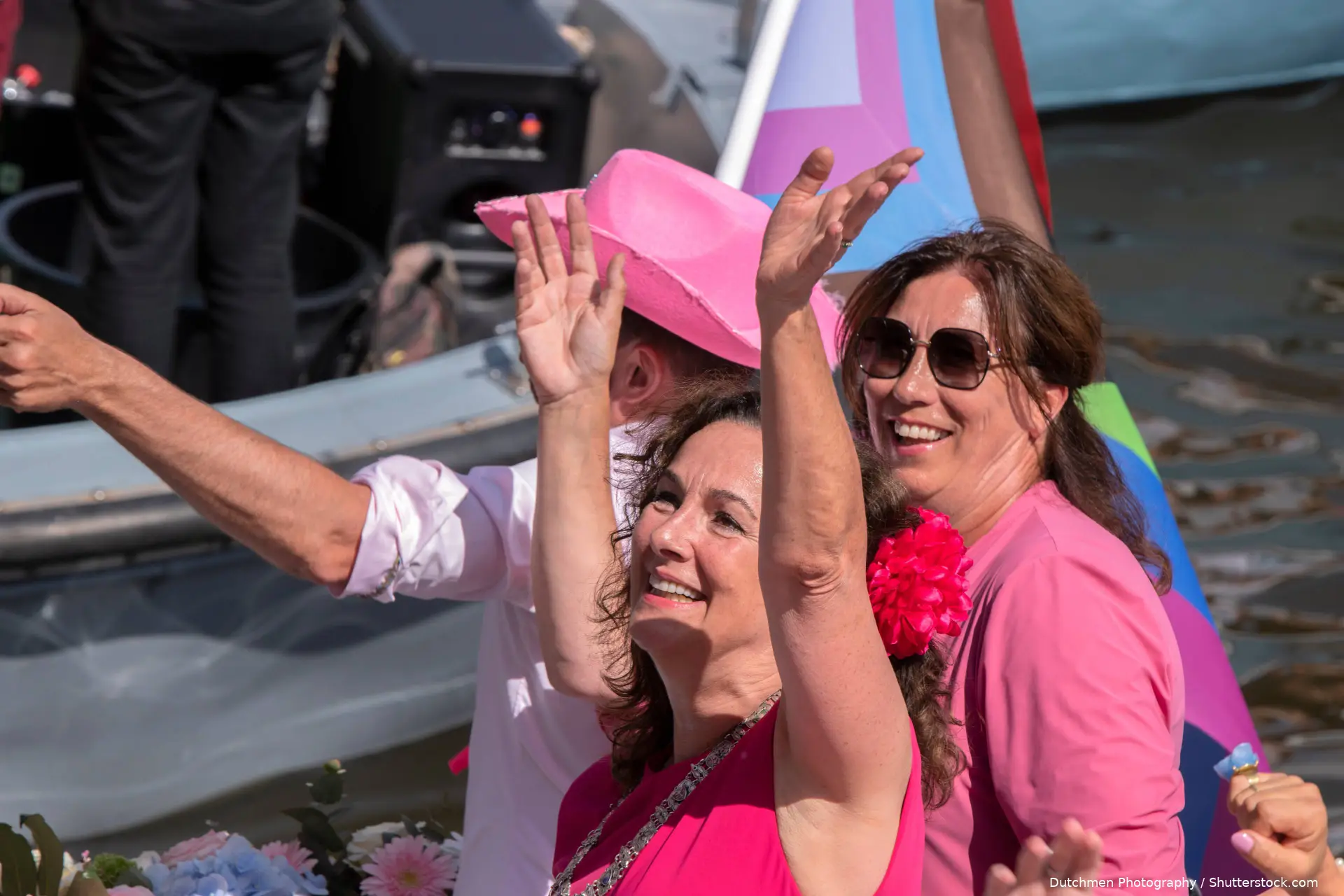 Femke Halsema zwaait lachend vanaf een boot met een regenboogvlag, in een roze shirt met een bloem in het haar