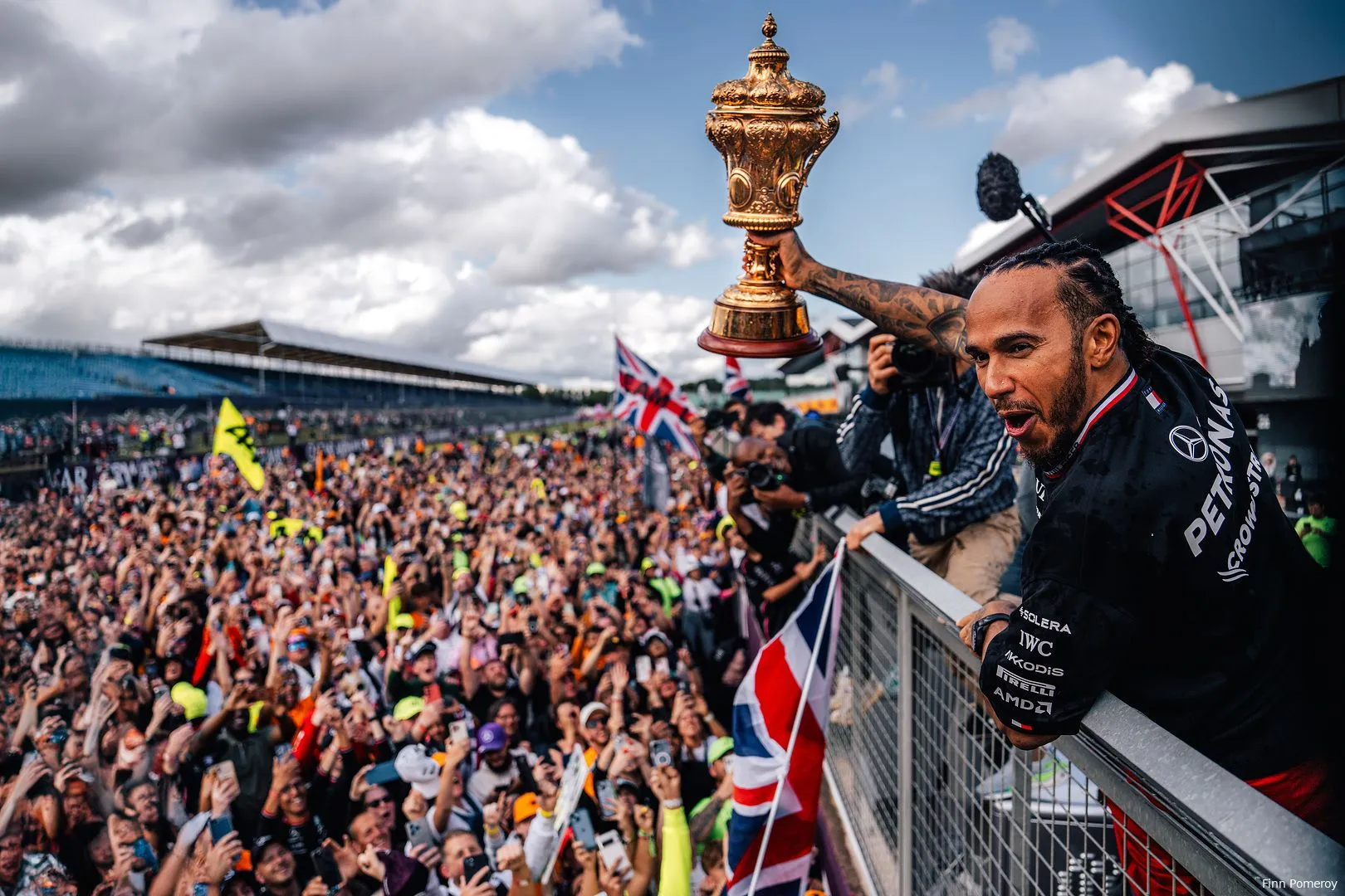 Lewis Hamilton celebrates with British fans on track at Silverstone Circuit after winning the 2024 British Grand Prix, raising the trophy above his head with one hand.