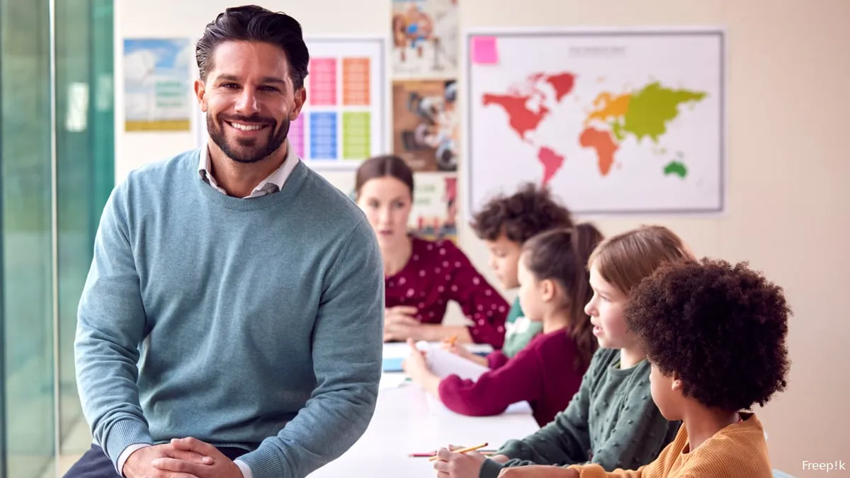 portrait smiling male elementary school teacher working desk classroom with students