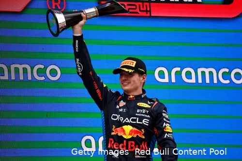 Max Verstappen smiling while holding the winner's trophy for the 2024 Spanish Grand Prix high above his head with his right hand.
