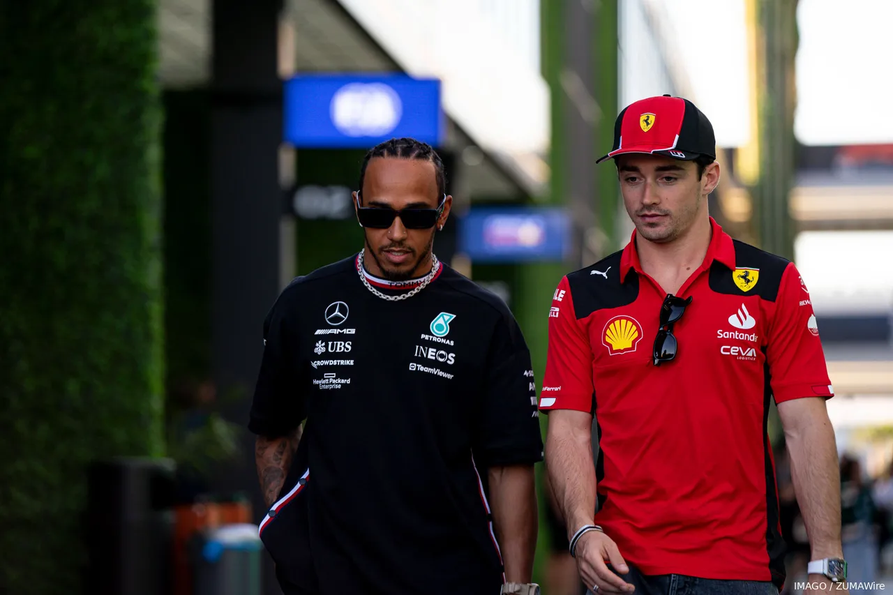 Lewis Hamilton and Charles Leclerc walking together through the paddock at one of the F1 circuits, with Red Bull hospitality and the FIA office in the background.