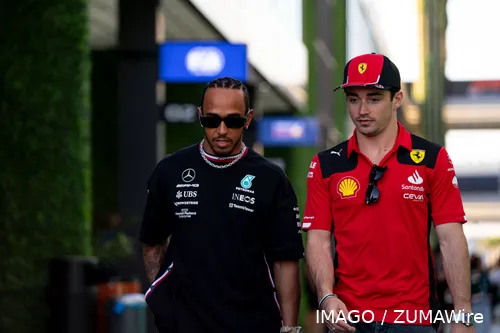 Lewis Hamilton and Charles Leclerc walking together through the paddock at one of the F1 circuits, with Red Bull hospitality and the FIA office in the background.