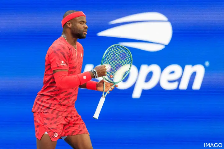 Tiafoe walks with the racket in his left hand and the US Open logo in the background