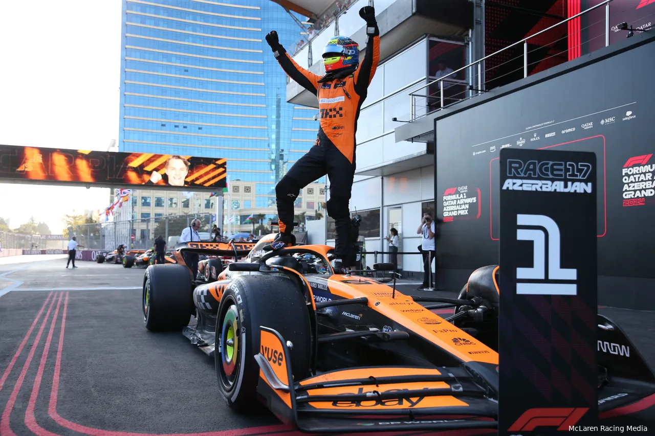 Oscar Piastri standing on top of his McLaren, both hands raised in celebration after winning the 2024 Azerbaijan Grand Prix, with Baku buildings in the background.