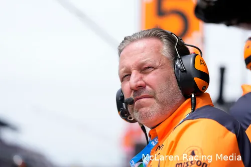 Zak Brown in McLaren team clothes, wearing a headset on the McLaren pit wall with a blurry background.