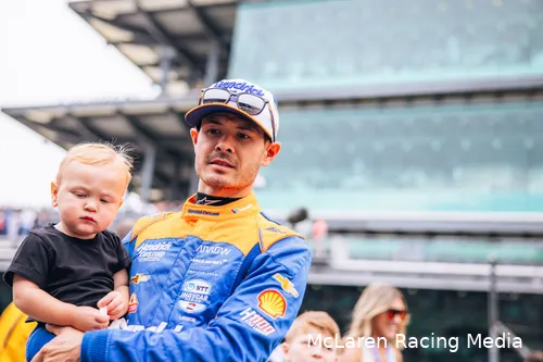 Kyle Larson in an Arrows McLaren racing suit, holding his child, with facility buildings of an IndyCar circuit in the background.