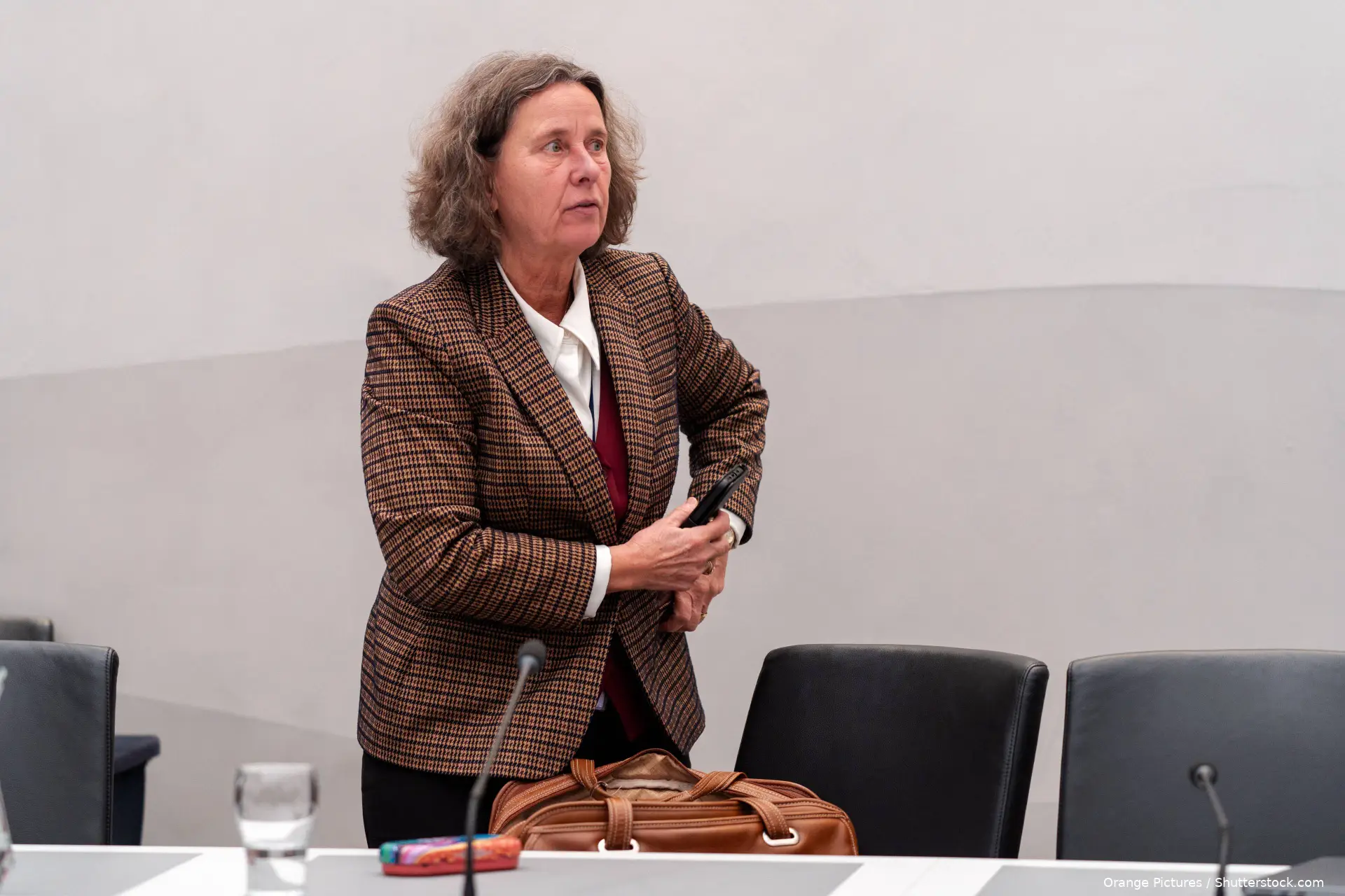 Marjolein Faber in een geruit colbert staat in een vergaderzaal en kijkt opzij