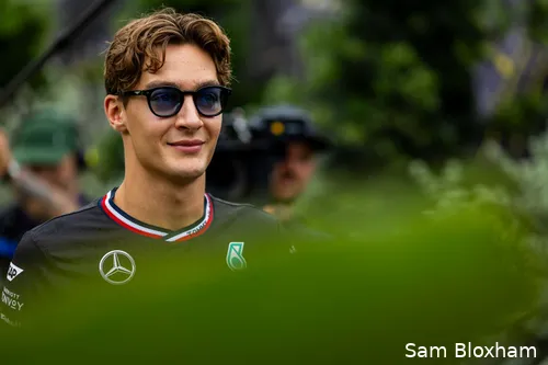 George Russell in Mercedes team clothes at the Singapore Grand Prix paddock, wearing glasses, with a camera crew in the background, shot from behind a green plant.