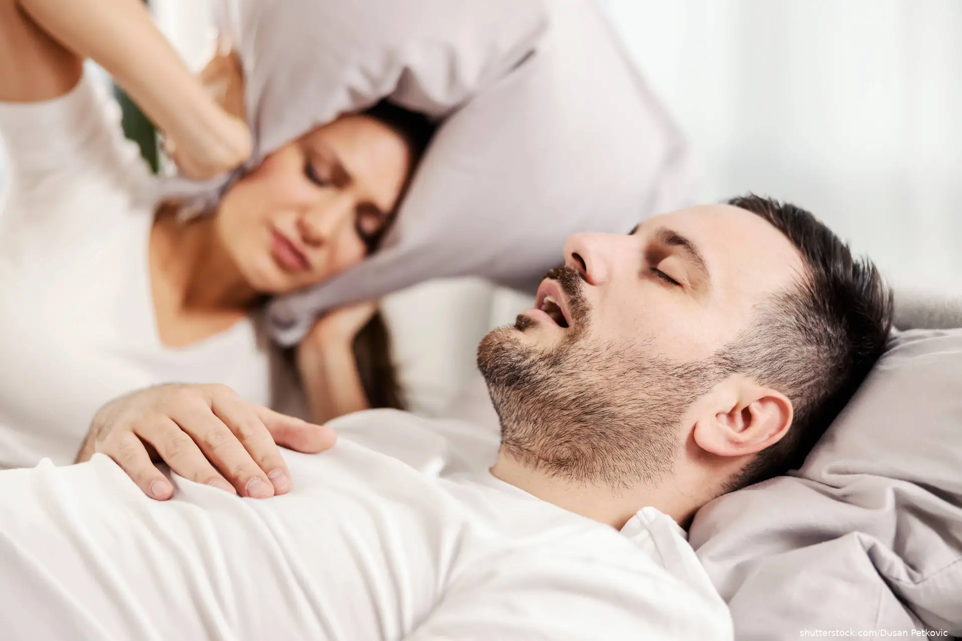 A man snoring and sleeping in the bed while woman is annoyed