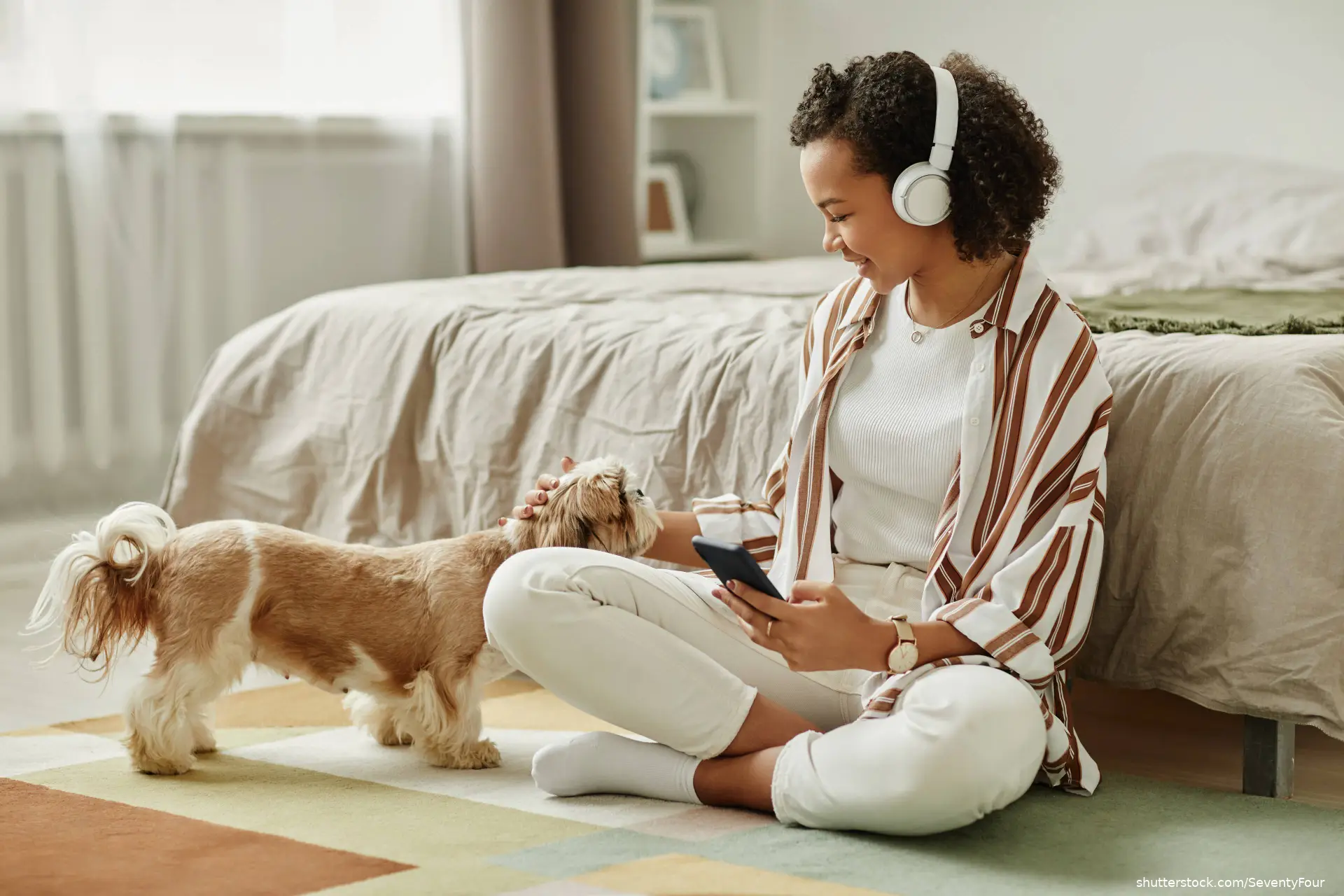 young woman playing with little dog while sitting on floor holding smartphone
