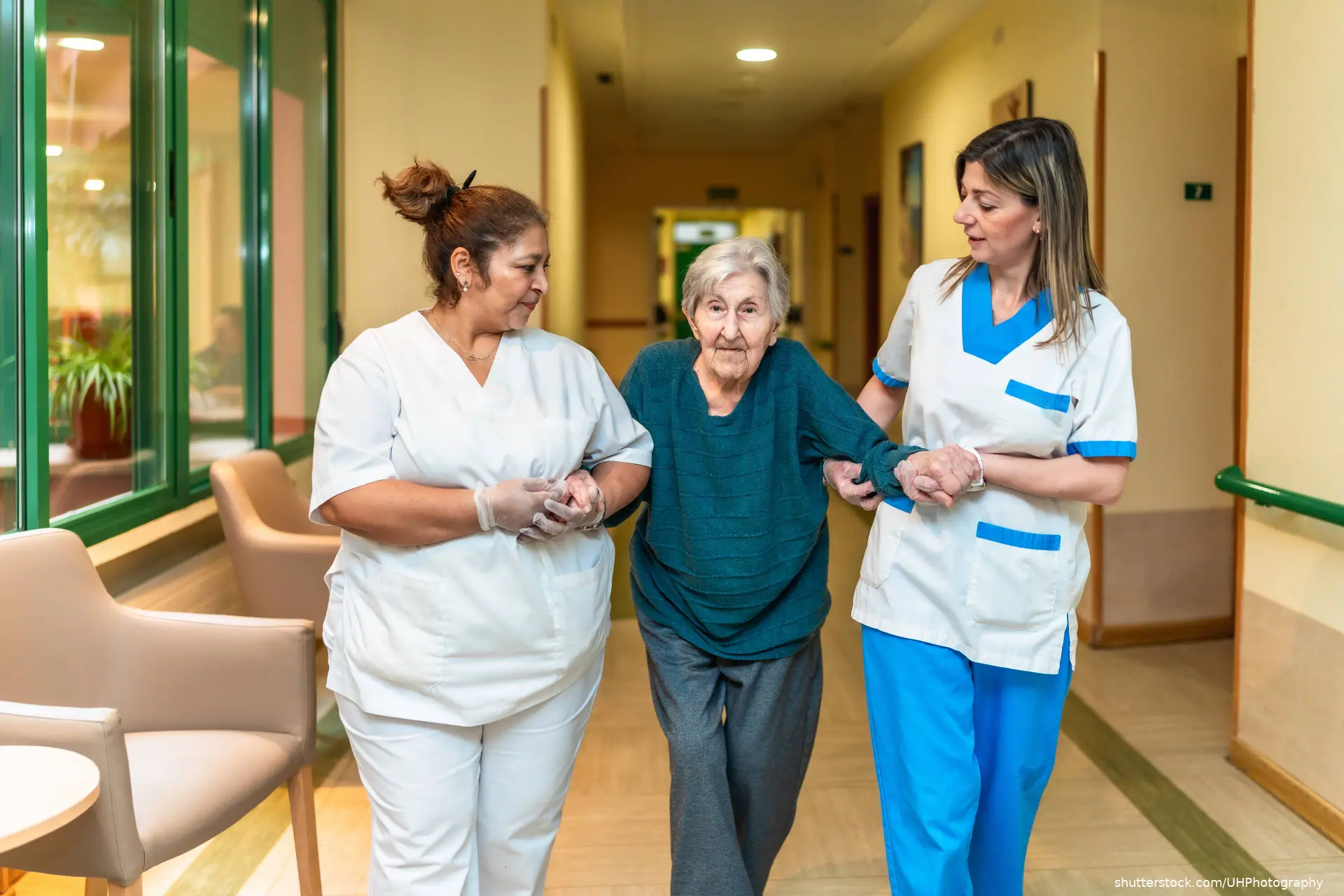 Two nurses assisting an elderly woman walking down the corridor shutterstock_2618843089